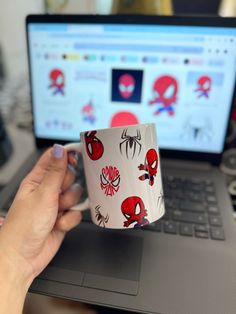 a person holding a coffee mug in front of a laptop computer with spiderman stickers on it