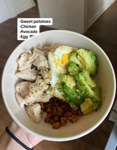 a person holding a white bowl filled with meat, vegetables and an egg on top