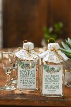 two glass jars with labels on them sitting on a table next to wine glasses and succulents