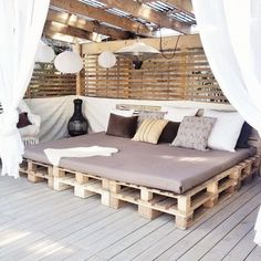 a bed made out of wooden pallets on top of a deck with white curtains