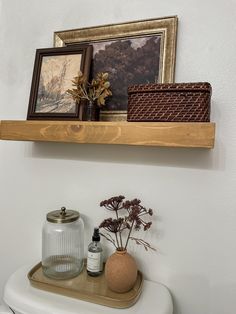 a white toilet sitting next to a wooden shelf with pictures above it and a vase filled with flowers