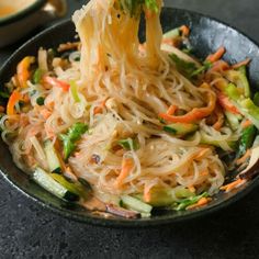 someone holding chopsticks with noodles and vegetables in a black bowl on a table