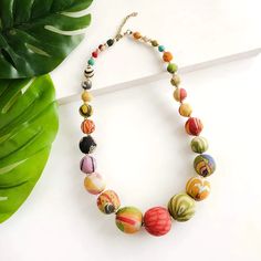 a multicolored beaded necklace sitting on top of a white table next to a green leaf