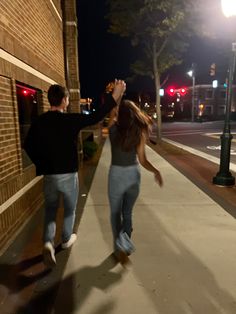 two people are walking down the sidewalk near a brick building at night, one person is reaching up into the sky