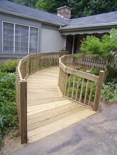 a wooden deck in front of a house with trees and bushes around it, next to a driveway