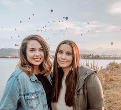 two women standing next to each other near the water with hot air balloons in the sky