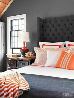 a bed with orange and white pillows in a bedroom next to a dresser, lamp and window