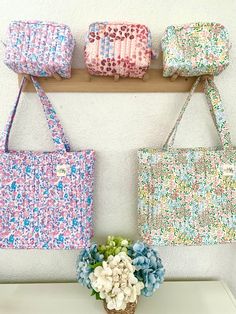 three purses hanging on a wall with flowers in the foreground and a vase full of hydrangeas behind them