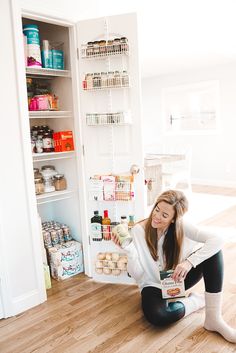 a woman sitting on the floor in front of an open pantry holding a box of food