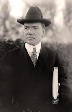 an old photo of a man in a suit and hat holding a book while standing next to a bush