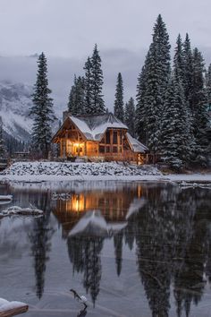 a log cabin is lit up at night by the water's edge with snow on the ground