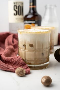 two glasses filled with liquid sitting on top of a table next to bottles and nuts