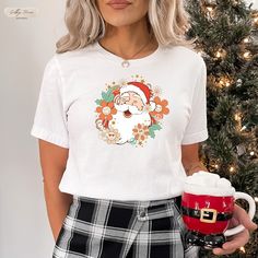 a woman wearing a santa clause t - shirt holding a cup of coffee in front of a christmas tree