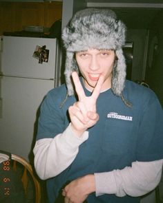 a young man wearing a hat making the peace sign with his hand while standing in front of a refrigerator