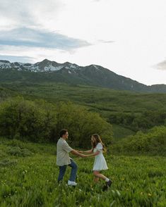 a man and woman are holding hands in the middle of a grassy field with mountains in the background