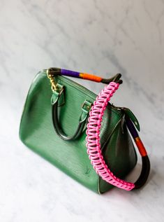 a green handbag with pink and purple braiding on the handle sits on a marble surface