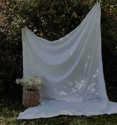 a white cloth draped over a basket with flowers in it on the ground next to some bushes