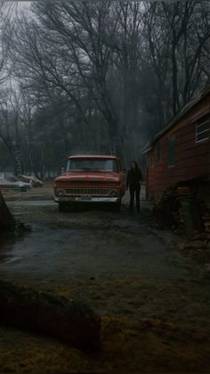 an old red truck parked in front of a house on a rainy day with two people standing next to it