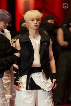 a man with blonde hair wearing white pants and a black jacket standing in front of other men