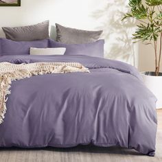 a bed with purple sheets and pillows in a room next to a potted plant