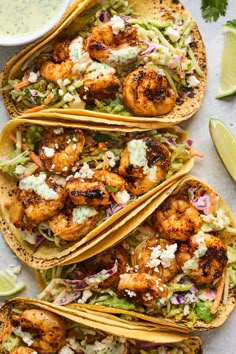 three tacos with shrimp, lettuce and cole slaw on them next to lime wedges