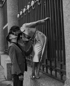 two women standing on the side of a street next to a fence and one is kissing the other