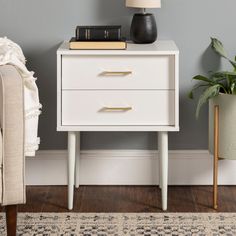 a white nightstand with two books on it next to a beige chair and potted plant