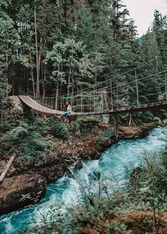 a man walking across a suspension bridge over a blue river in the middle of a forest