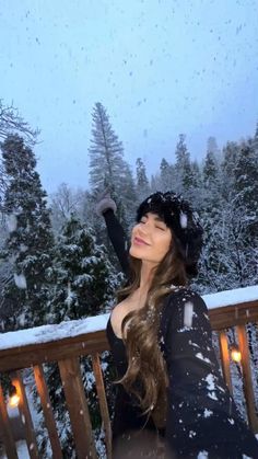 a beautiful woman standing on top of a balcony next to snow covered trees and holding her arms up in the air