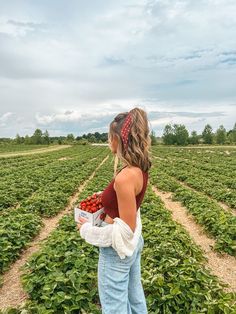 a woman standing in a field holding a box of strawberries