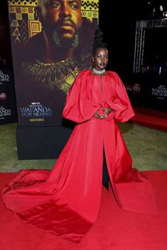 a woman in a red gown standing on a red carpet with a poster behind her