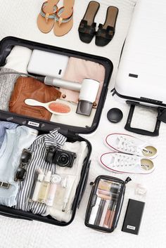 an open suitcase filled with clothes, shoes and other items sitting on a bed next to a pair of flip flops