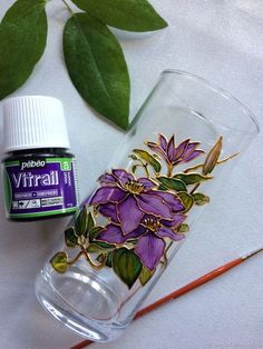 a glass filled with purple flowers next to a paintbrush and some green leafy leaves