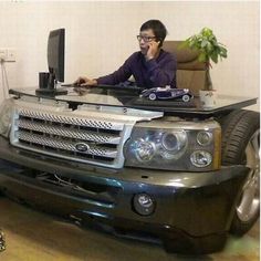 a man sitting in front of a computer on top of a black car with the hood up