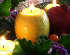 two oranges and some candles sitting on a table with green leaves around them, surrounded by purple flowers