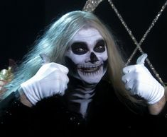 a man with long hair and white makeup wearing skeleton makeup, holding two hands up in front of his face