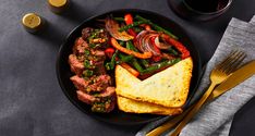 a black plate topped with meat and veggies next to two glasses of wine