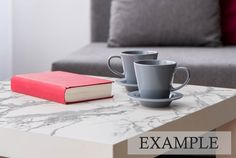 two coffee cups sitting on top of a marble table next to a red book and gray couch