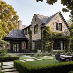 a large stone house surrounded by greenery and trees with an outdoor dining table in the foreground