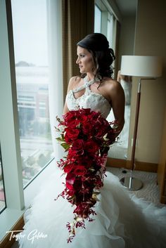 a woman in a wedding dress holding a bouquet of red flowers looking out the window