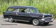 an old black car parked in a parking lot next to some tall green grass and bushes