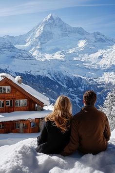 a man and woman sitting on top of a snow covered hill looking at the mountains