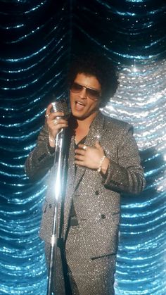 a man in a suit and sunglasses holding a microphone while standing next to a curtain