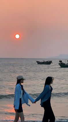 two women walking on the beach holding hands with boats in the water behind them at sunset