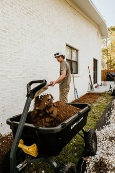 a man is pushing a wheelbarrow filled with dirt and mulch in front of a house