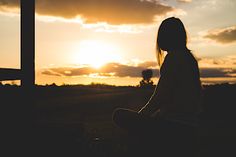 a woman sitting on the ground watching the sun go down in the distance with her hands behind her back