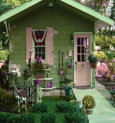 a small green house with pink shutters and potted plants on the front porch