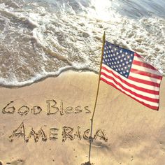 an american flag is on the beach with words written in sand and waves behind it