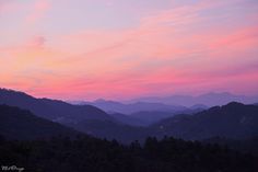 the sky is pink and purple as the sun sets in the distance over mountains with trees