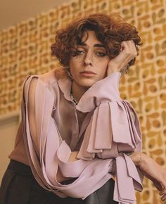 a woman with curly hair wearing a pink blouse and black pants, holding her hand on her head